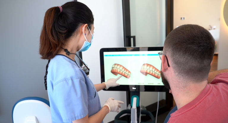 A dental professional in a blue uniform and face mask points to a computer screen displaying two 3D tooth models during a consultation. The patient, wearing glasses and a red shirt, sits attentively beside the desk, viewing the models. The image depicts a modern dental office, highlighting personalized care and digital imaging technology used to explain treatment options.
