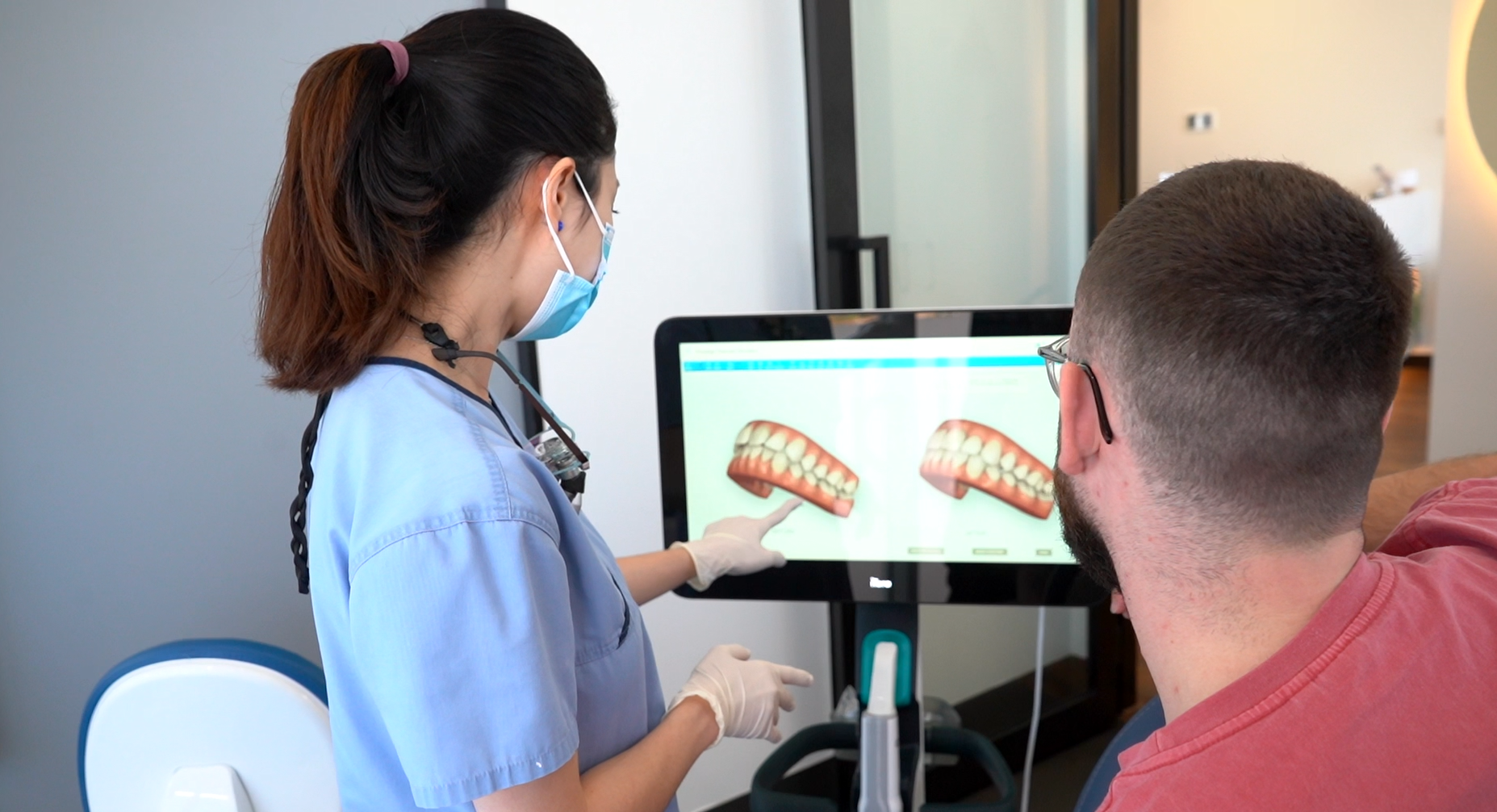 A dental professional in a blue uniform and face mask points to a computer screen displaying two 3D tooth models during a consultation. The patient, wearing glasses and a red shirt, sits attentively beside the desk, viewing the models. The image depicts a modern dental office, highlighting personalized care and digital imaging technology used to explain treatment options.