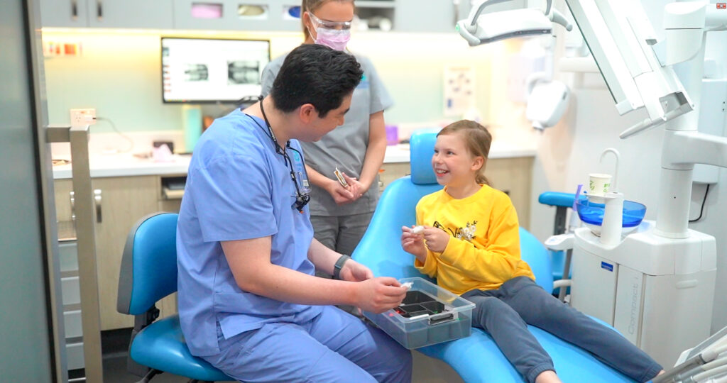 A young child sits in a dental chair, curiously engaging with a dental tool while a dental professional in blue scrubs explains something using another instrument. A second team member in grey scrubs stands nearby, smiling. In the background, a monitor displays dental X-rays, and the clinic is equipped with standard dental instruments and overhead lighting. The scene conveys a warm, educational, and professional environment, highlighting child-friendly care and teamwork in a modern dental practice.