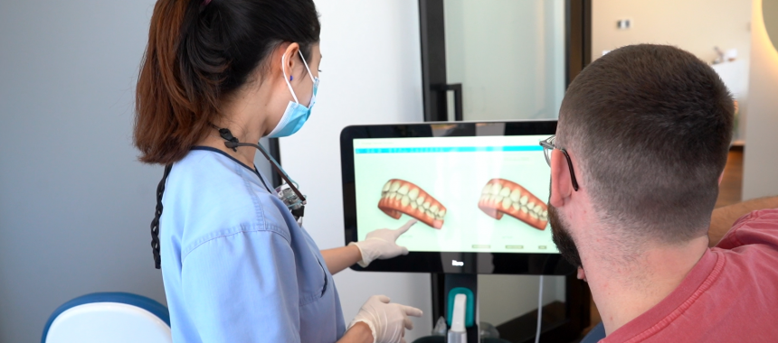 A dental professional in a blue uniform and face mask points to a computer screen displaying two 3D tooth models during a consultation. The patient, wearing glasses and a red shirt, sits attentively beside the desk, viewing the models. The image depicts a modern dental office, highlighting personalized care and digital imaging technology used to explain treatment options.